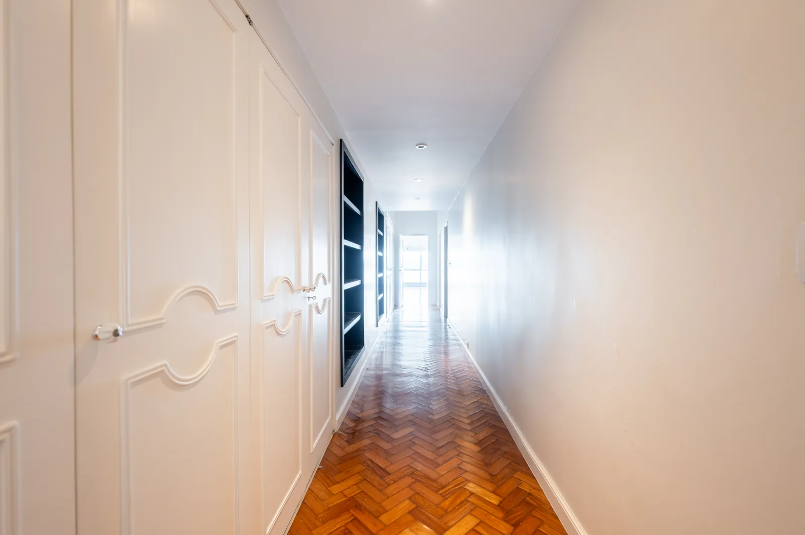 A hallway lined with white fitted cabinetry over herringbone parquet