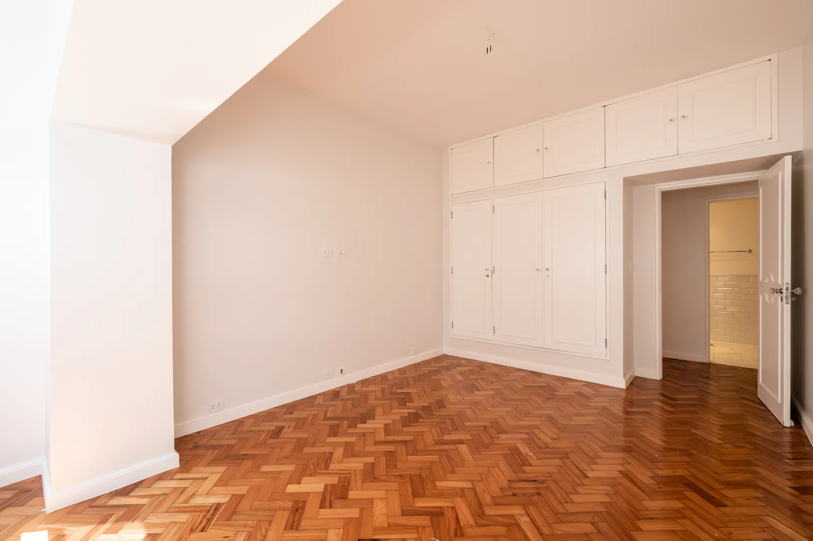 A bedroom with white built-ins, parquet and a doorway through